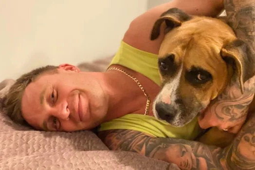 A man with tattoos with a yellow singlet holds a dog in bed.