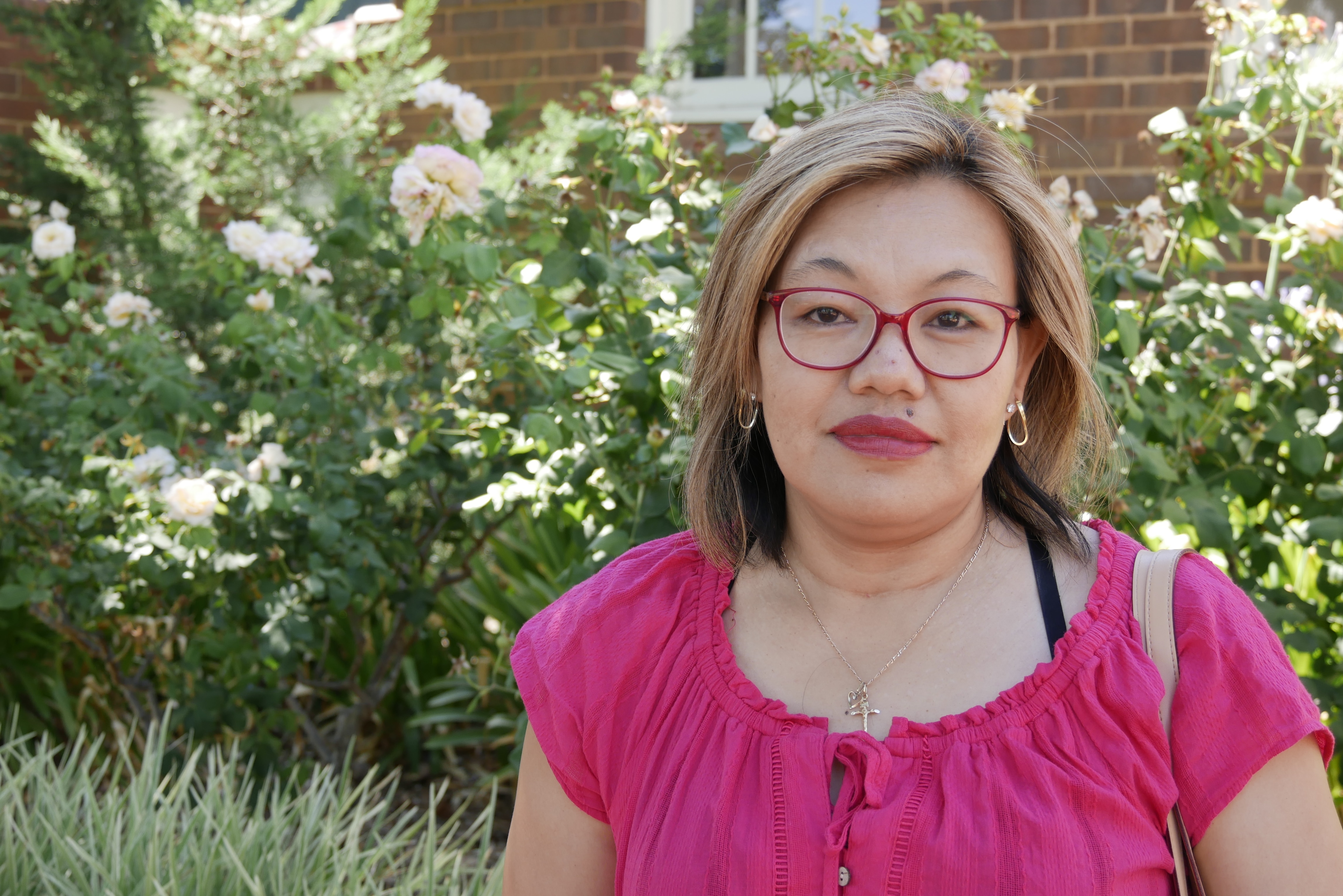 Una mujer de cabello rubio y camisa rosa con gafas se para frente a un rosal y no sonríe. 