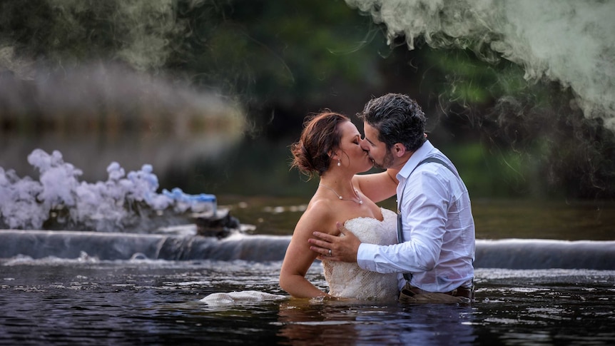 A man and a woman wearing wedding attire stand in a creek.