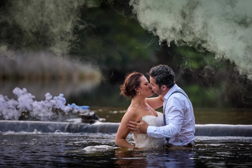 A man and a woman wearing wedding attire stand in a creek.