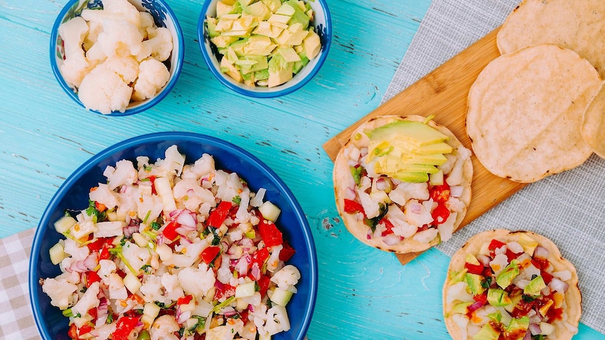 Brightly coloured cooked cauliflower with herbs in blue bowl surrounded by other ingredients in bowls and tostadas on a table.