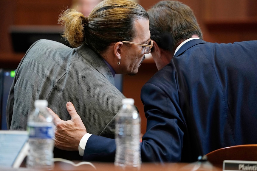 A man with a grey suit whispers into the ear of a man in a dark blue suit whose hand rests on the former's back