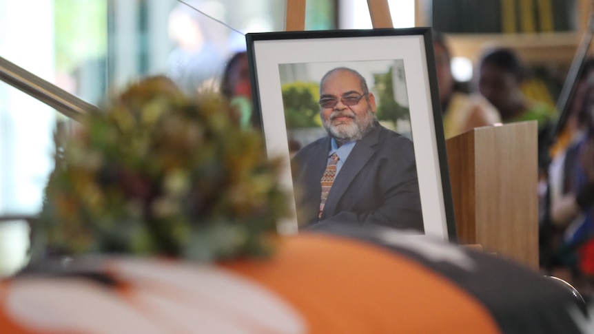 A framed photo of a man atop a casket drapped in the Northern Territory flag, inside a church.