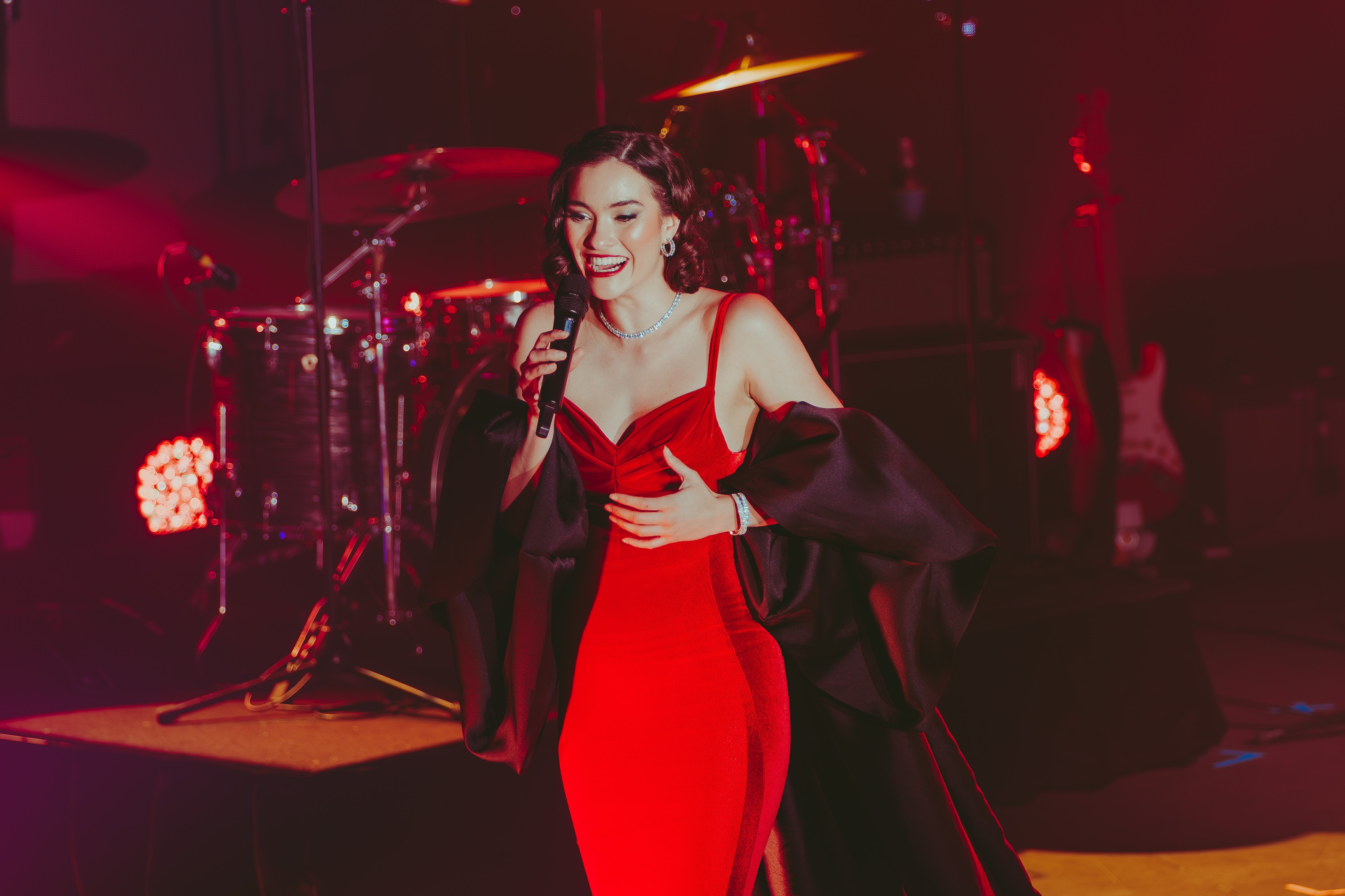 Nina Korbe wears a red velvet formal gown with a black silk cape and sings into a microphone on stage with a broad smile.