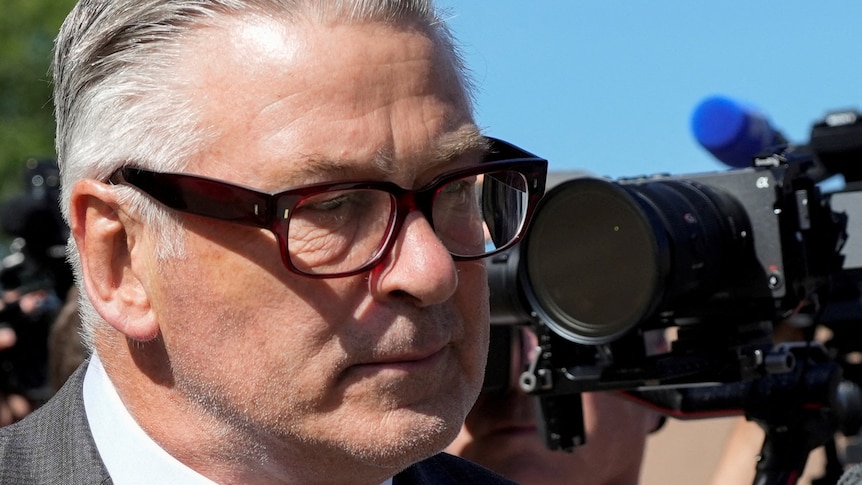 A close-up of Alec Baldwin's face, looking solemn and away from camera, wearing thick glasses, grey hair