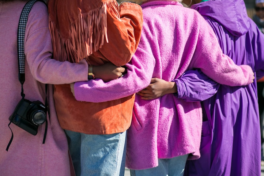 Backs of women, wearing pink and purples jackets, with arms wrapped around each other.