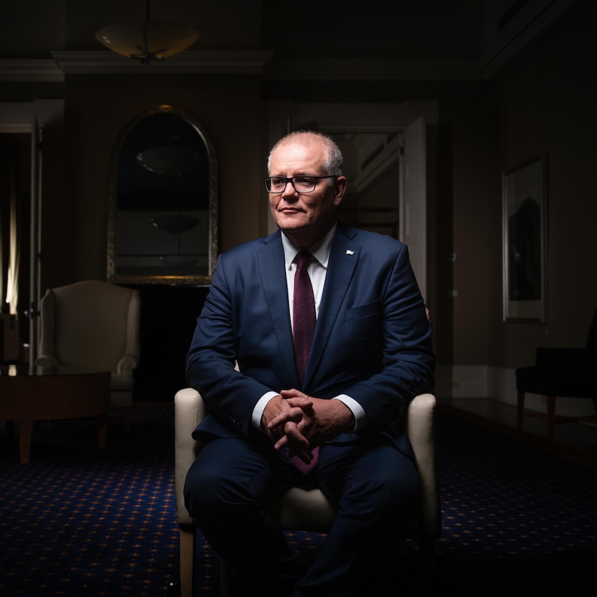 Dressed in a blue suit, Scott Morrison sits in a chair in a large room, looking to the side.