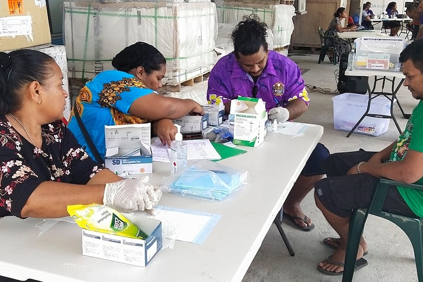 Several people sit around a portable table covered in paperwork and medical paraphernalia.