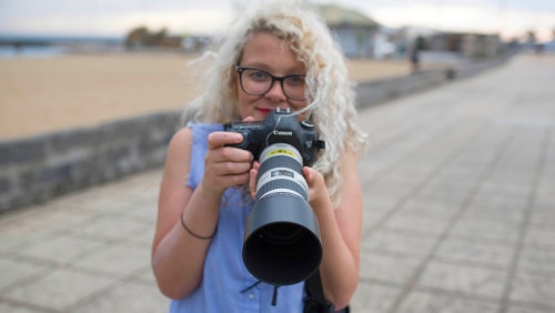 Margaret smiles as she holds a camera with a long lens up to her face.