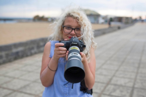 Margaret Burin holding camera with long lens looking to camera with beach background.