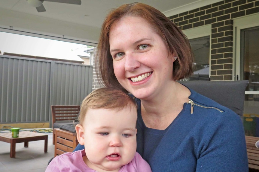 A mother holds her baby on her lap, in front of a brink and tile home. She's smiling.