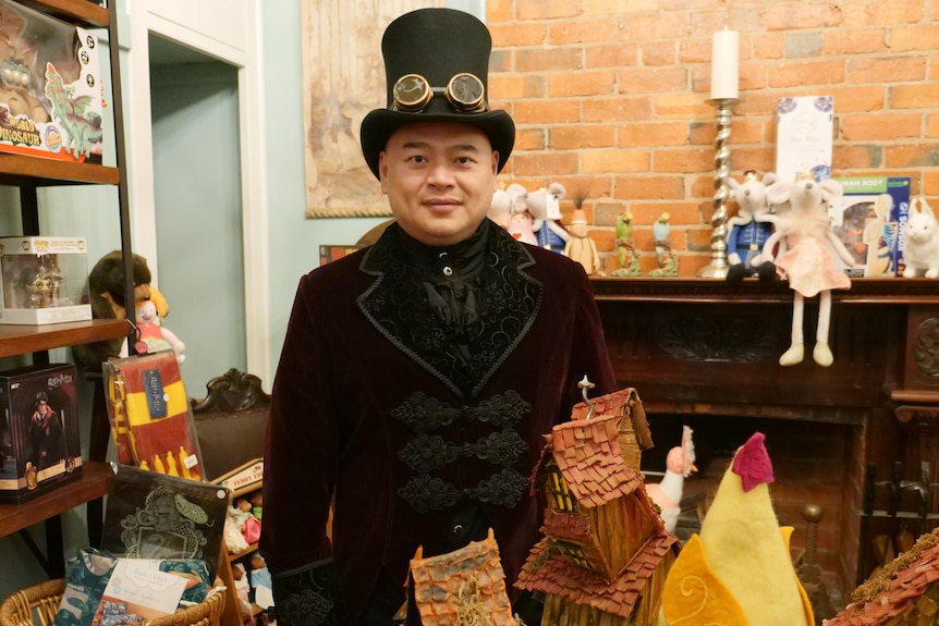 A man in a top hat and vintage jacket poses with antiques in his shop.