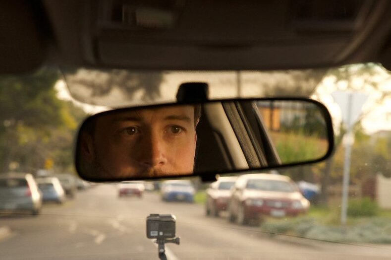 A young man's face is reflected in the rearview mirror as he drives a car through a suburban street in the daytime.