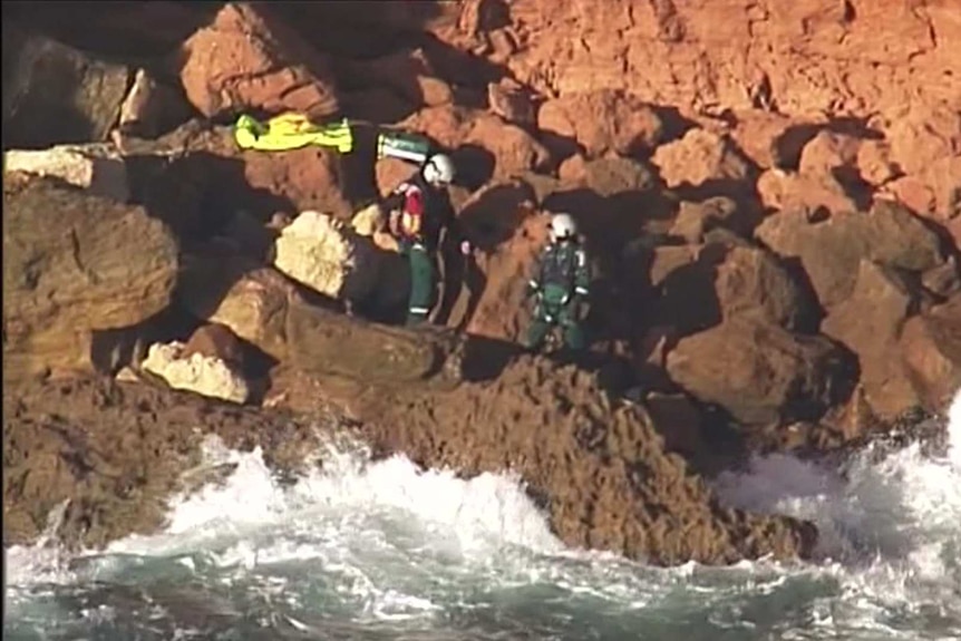 Two emergency service officers wearing green overalls stand at the base of a rocky cliff