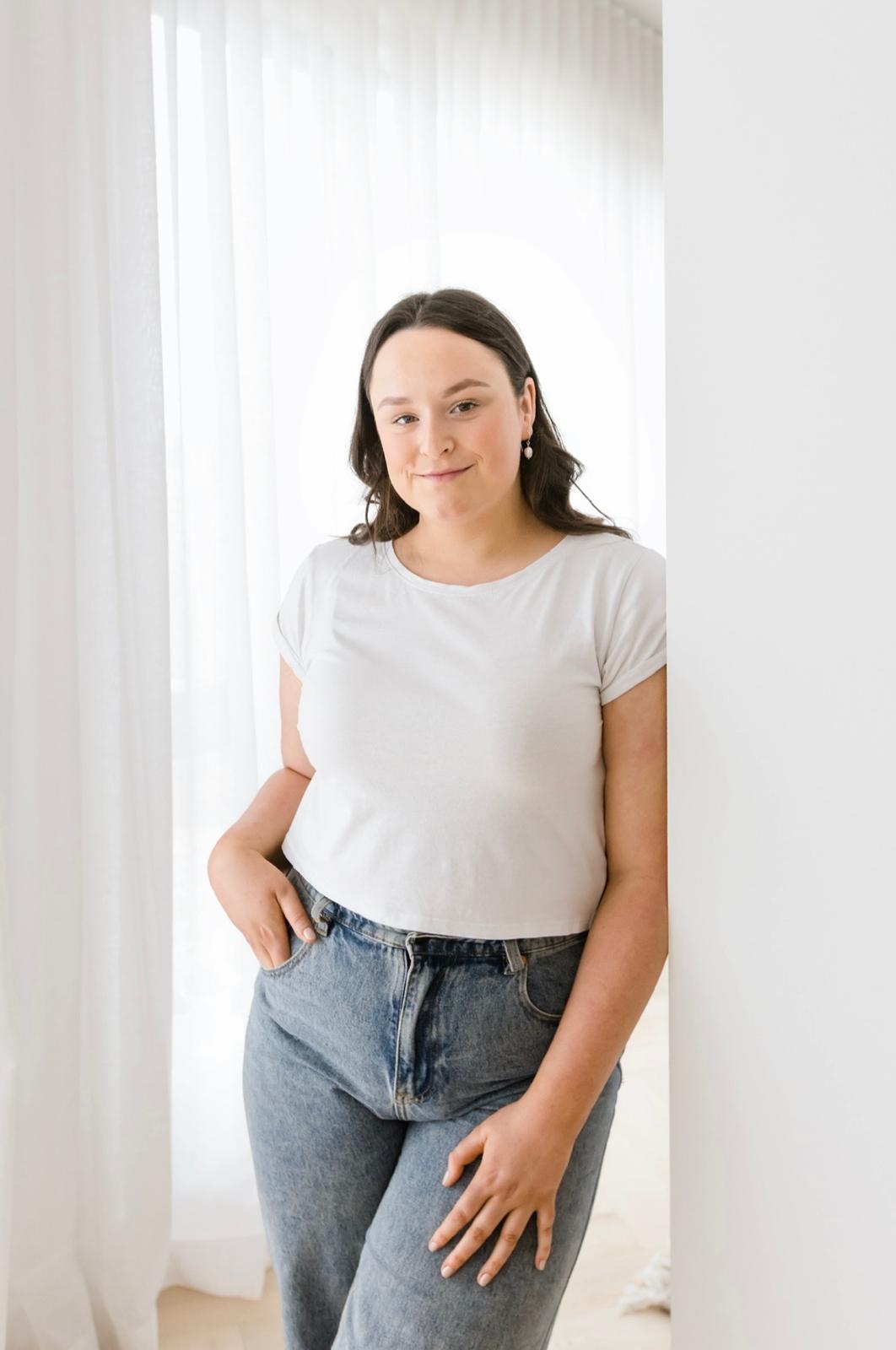 A young woman with brown hair wearing a white top and blue jeans leans against a wall, smiling