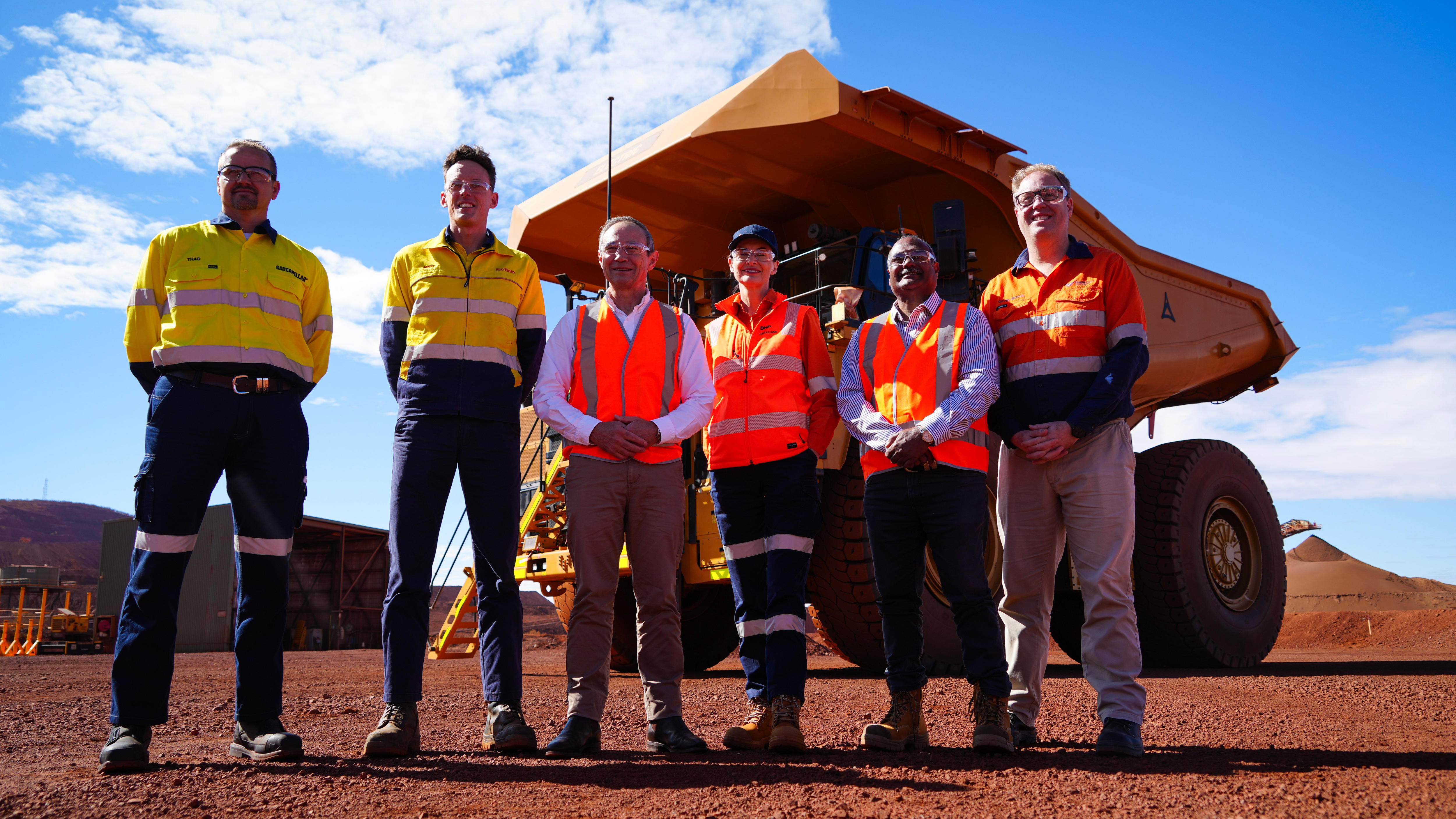 A group in high-visibility stand at attention with a giant truck behind them.