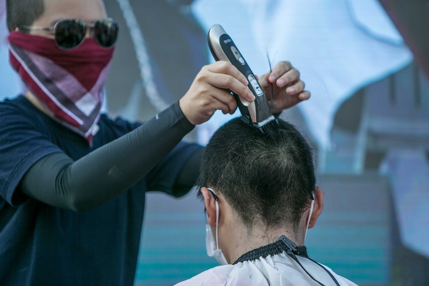 A woman in sunglasses and balaclava shaving another woman's head.