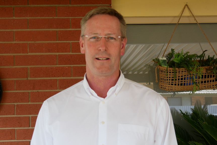 A man with light hair and glasses and white shirt stands against a brick wall with a dartboard and plants in the background.