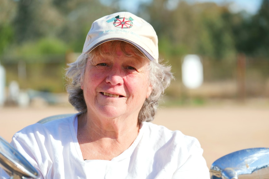A woman in a white top a baseball cap looks at the camera