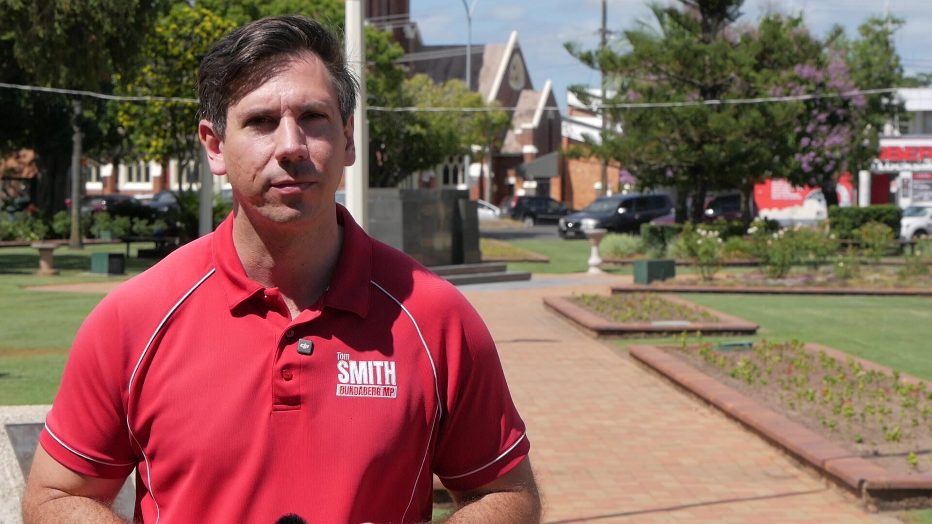 Un hombre con camisa roja y pelo negro se encuentra en un parque.