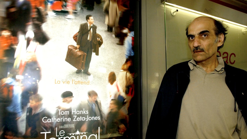Merhan Karimi Nasseri stands next to a poster for The Terminal starring Tom Hanks 