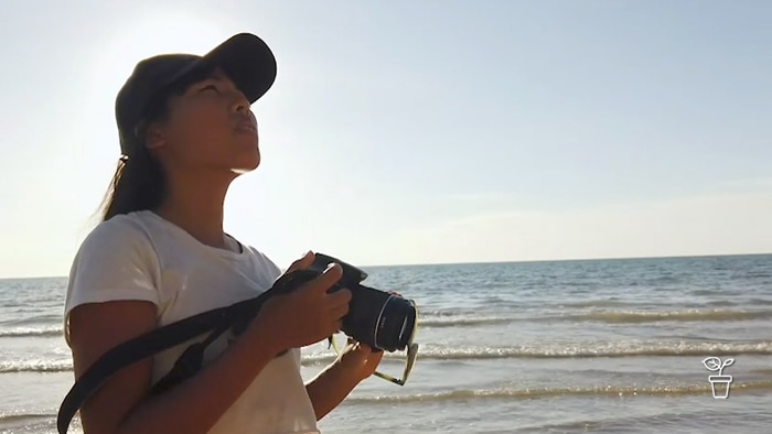 Girl wearing a hat standing on a beach holding a camera