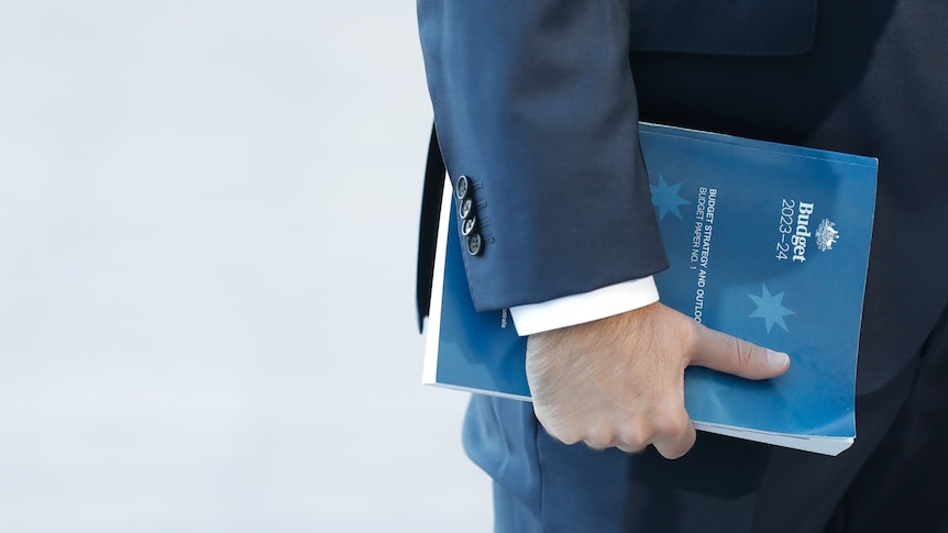 A close up of Jim Chalmers holding a thick book with a dark blue cover which a lighter blue Southern Cross titled Budget 2023-24