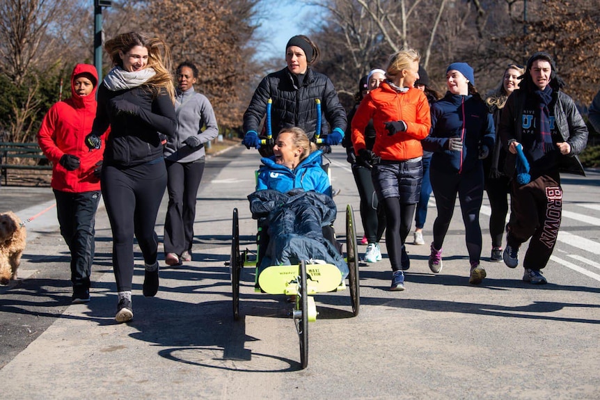Mina Guli pushed in a wheelchair through a park at night by 15 runners with skyscrapers in the background