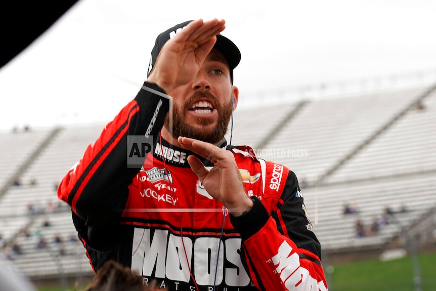 A racing car driver wearing red and black gestures while explaining something to crew members