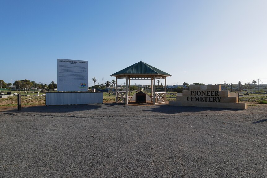 A large billboard with tourist information stands next to a small pavillion and sandstone sign reading 'Pioneer Cemetery'