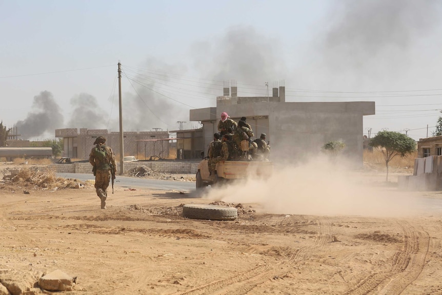 A truck packed with Turkey-backed Syrian fighters drives on a dirt road as smoke rises in the background.