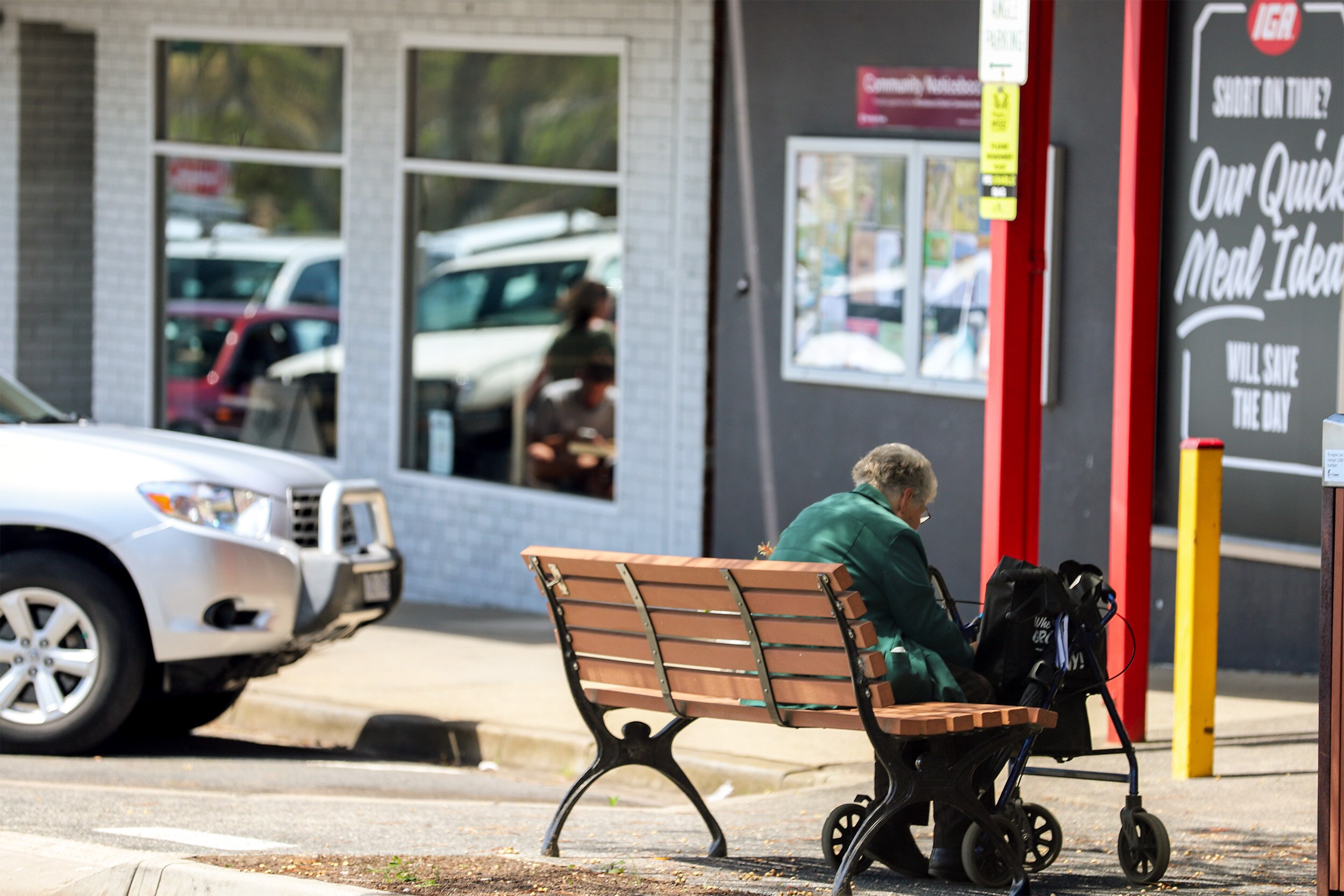 An older woman sits on a street bench with a walker in front of local shops
