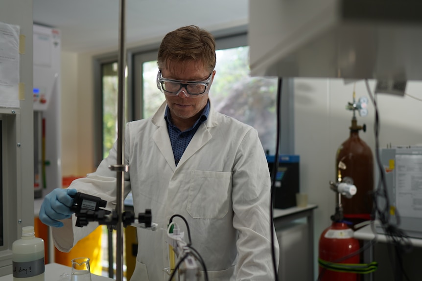 Man in a white lab coat with goggles and gloves conducting an experiment in a laboratory.