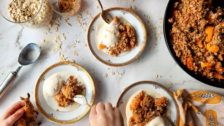 Birdseye view of a white table with three bowls of peach crumble served with vanilla ice cream. The crumble dish is on the right