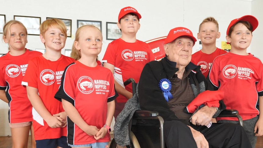 An elderly man in a wheelchair surrounded by six kids wearing red t-shirts that read 'Nambucca Nippers'
