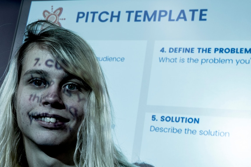 A young person of no gender stands in front of a projection of the words "define the problem".