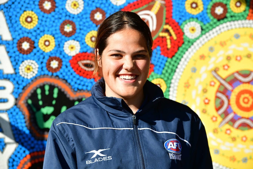 A young Indigenous woman in front of a colourful painted background with Aboriginal artwork on it.