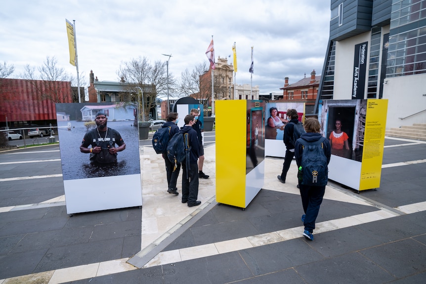 A group of teenage boys in school uniform stand looking at large photo boards in a public square.