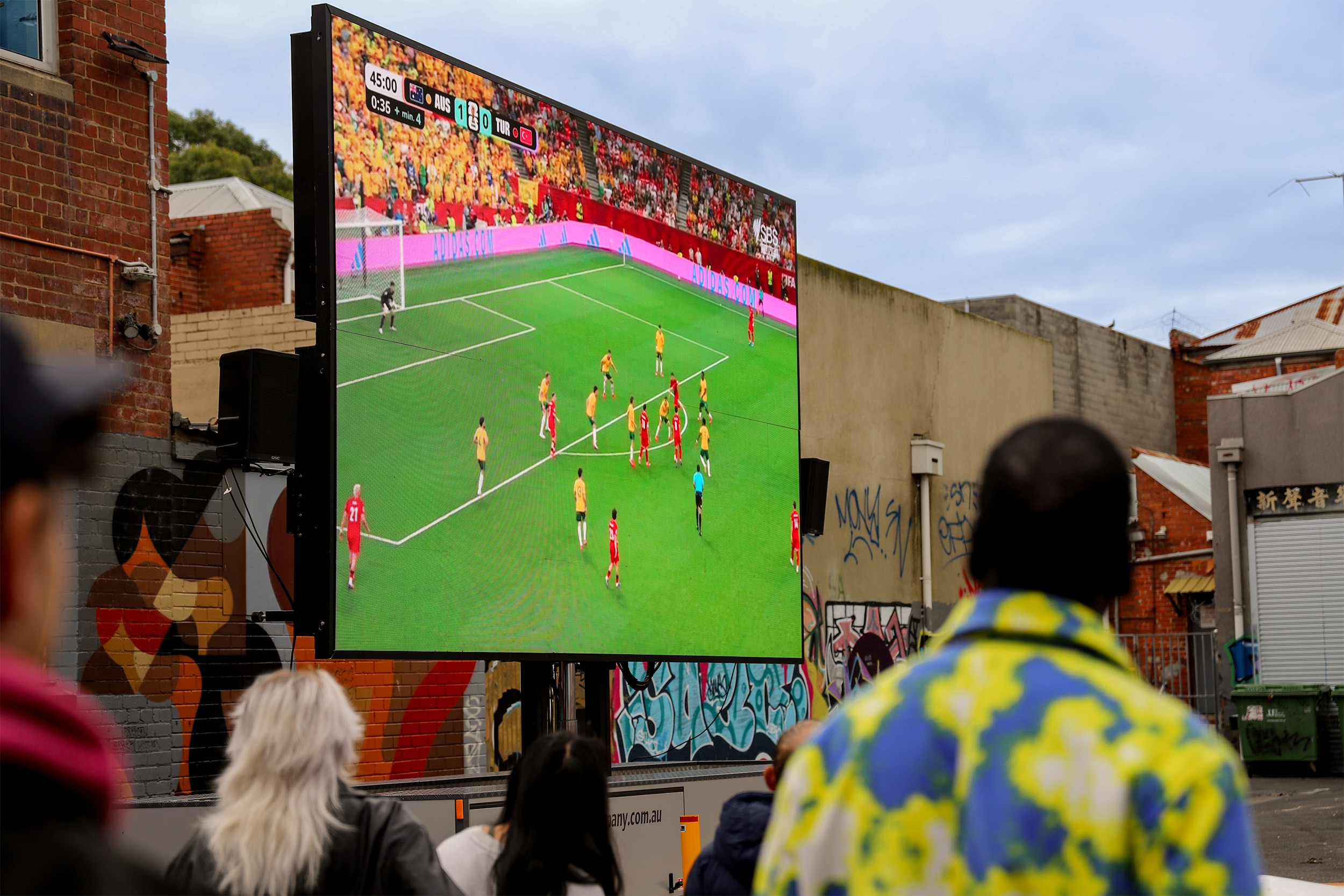 A large screen, showing a soccer pitch with players in the background, people can be seen in the foreground looking up at it.