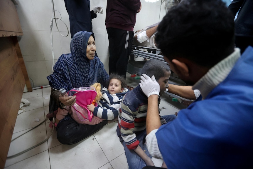 A woman sits on the floor holding a child while a man tends to another child at a hospital.