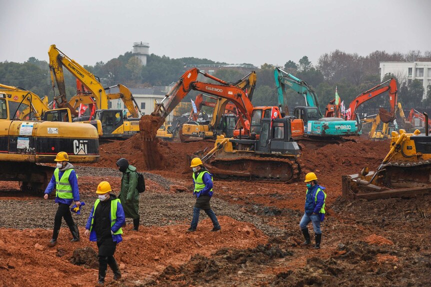 Five people walk to the left of the shot over open ground with several diggers of various colours behind them. The sky is grey.