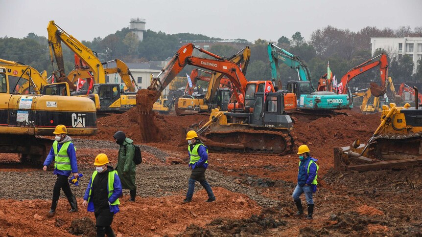 Five people walk to the left of the shot over open ground with several diggers of various colours behind them. The sky is grey.