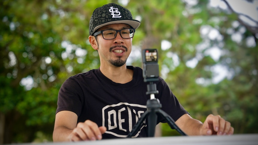 A man wearing cap and black Tshirt sits at a table with a small camera on a tripod in front of him