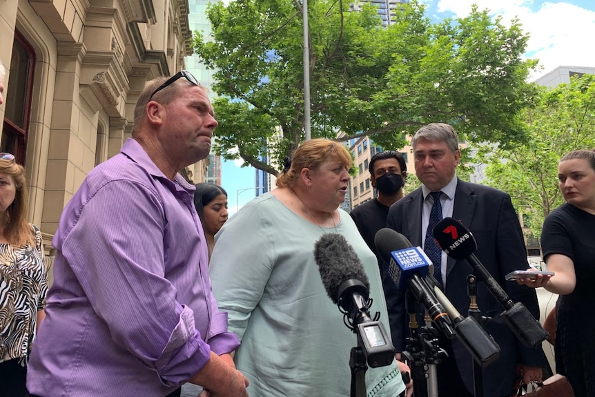 Three people standing outside a building with microphones in front of them.