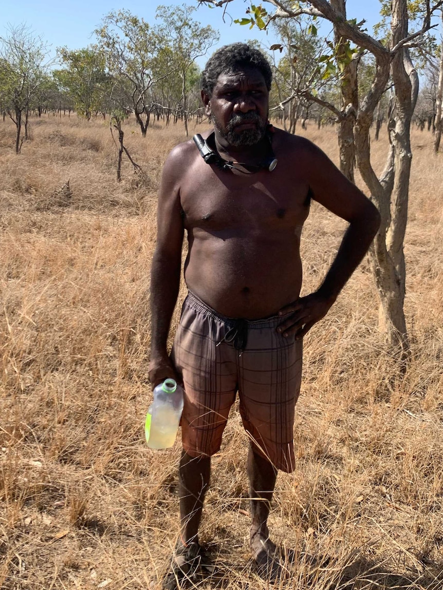 Aboriginal man stands in dry grassland on bright day with shorts on and holding near-empty drink bottle.