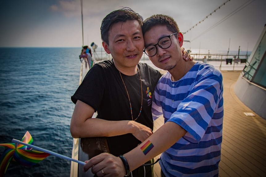 Two men stand smiling with their arms around each other on the deck of a cruise ship.