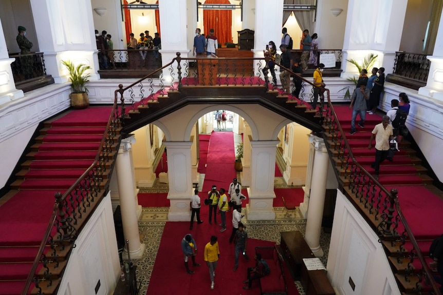 A shot looking down on an opulent, red-carpted stair case with people milling about on the ground floor