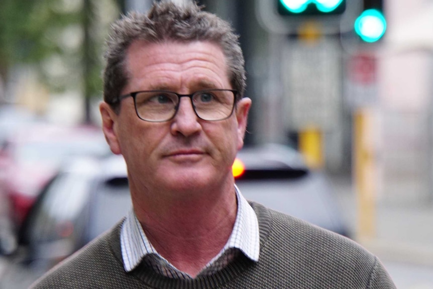 A man in glasses and a grey jumper walks down a street.