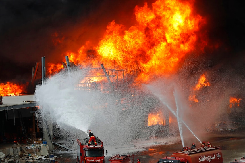 You view a massive fireball engulfing charred industrial structures with three firefighting vehicles looking miniscule below.