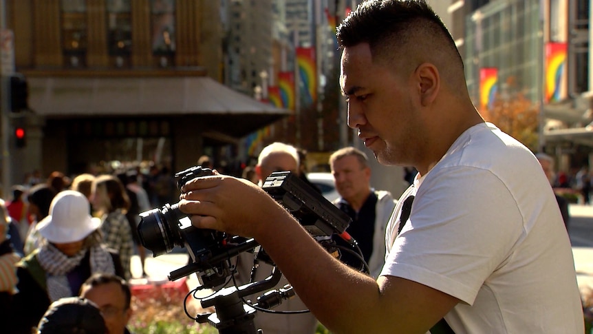 A man looks at a camera as he films in the city, surrounded by people.